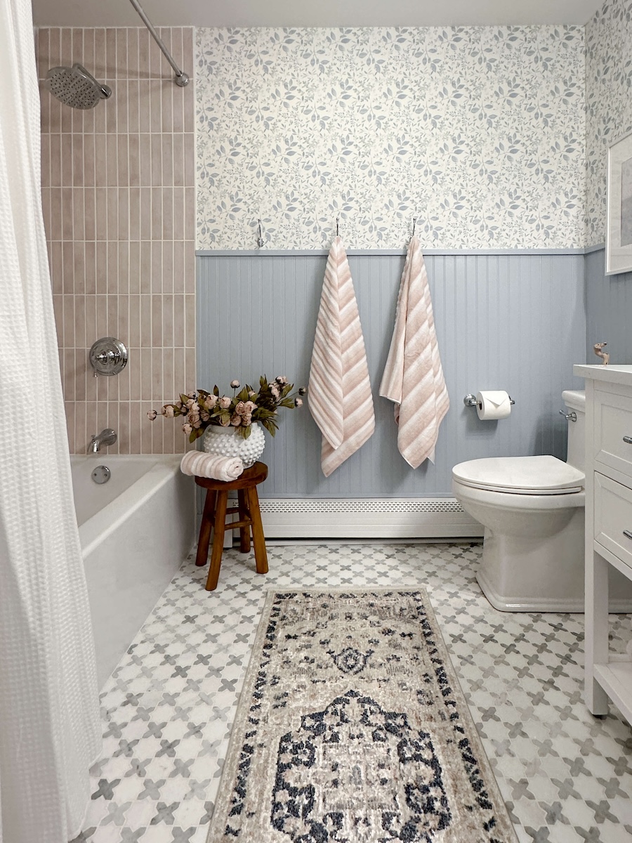 DIY Bathroom Reveal: Hanging blush towels on hooks with bluebead board and wallpaper on the walls. A quick glimpse of the finished tub with blush tiles, white shower curtain, and brand new tub with faucets. Brand new toilet installed on top of marbled geometric tiles. A pot of blush roses sit on a brown stool next to a rolled up towel. 