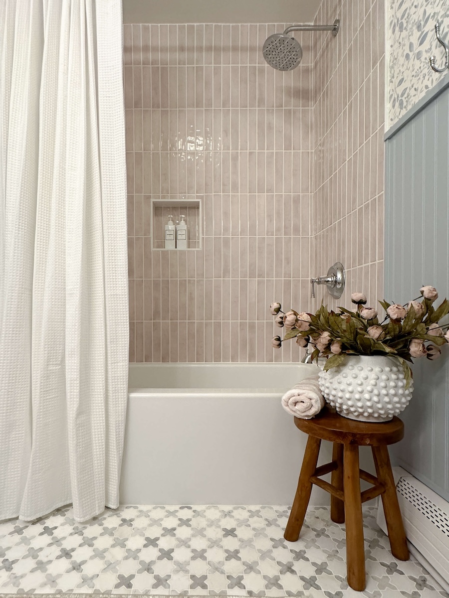 White geometric tile flooring and a stool with a white bowl that has bluhs roses sticking out of it sitting next to the brand new tub and white shower curtain from the DIY bathroom renovation. 