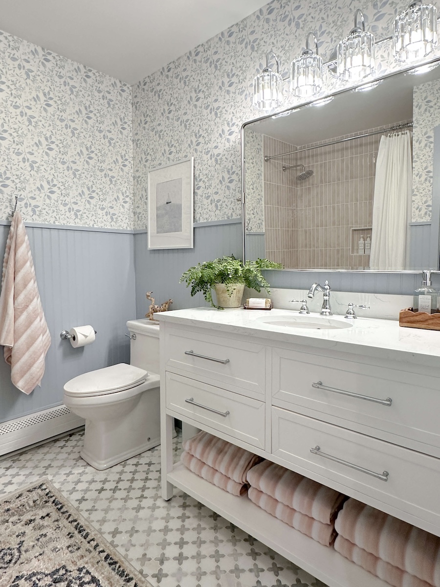 DIY Bathroom Renovation Reveal: White vanity with silver faucet and marble countertops. Hanging over the vanity is a silver framed mirror and lighting fixture. Blue wallpaper and blue beadboard on all the walls. White toilet and pink blush towels. 