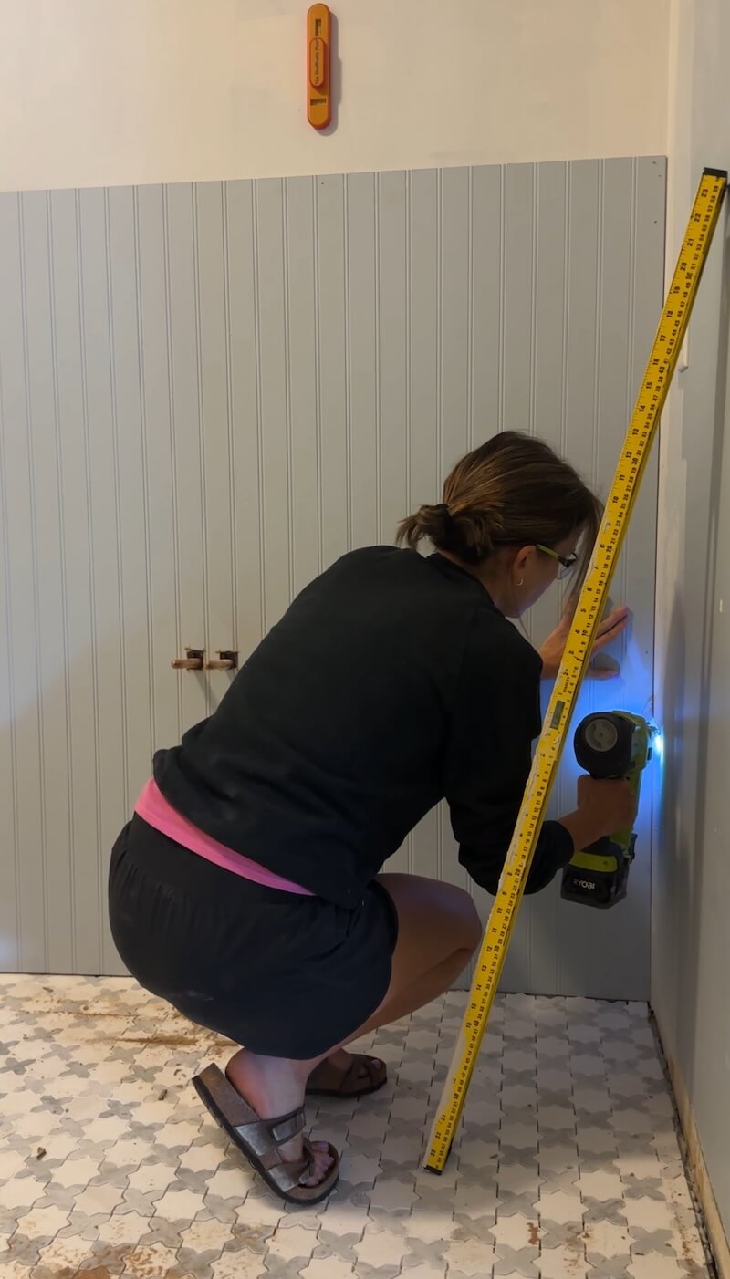 A woman crouching down with a nail gun to install the beadboard to the wall using nails in the corner. 