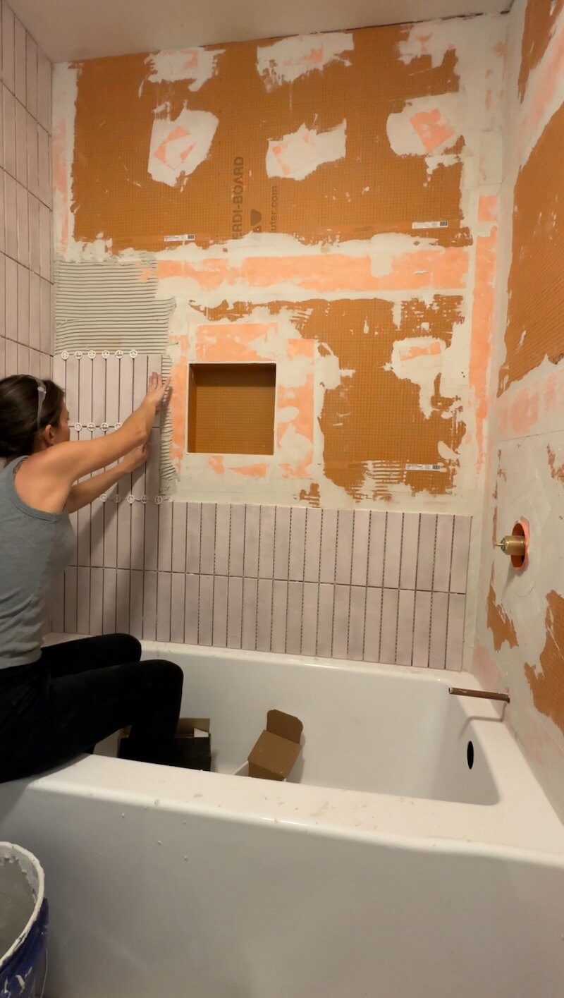 Melissa Tyler sitting on the edge of the tub pushing a tile onto the grout. 