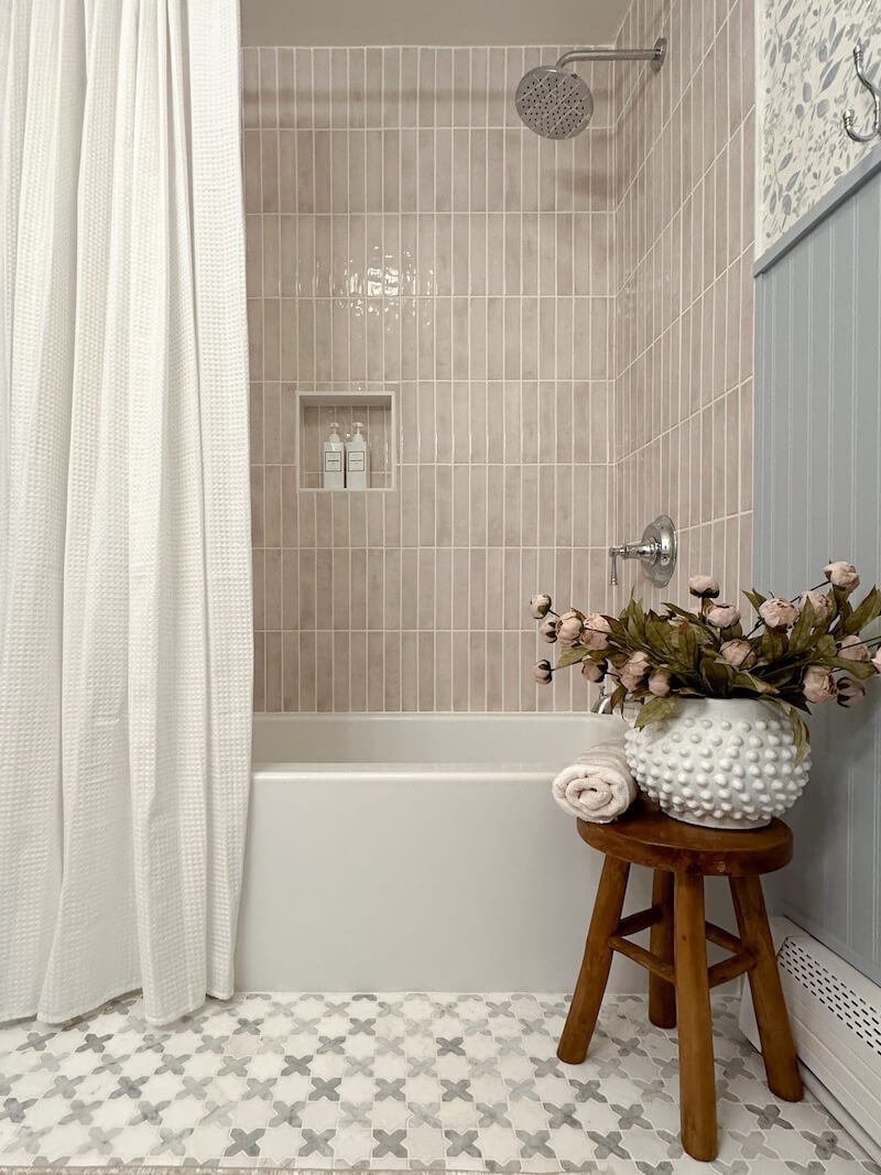 Final shower reveal with pink vertical tile, shower niche with shampoo, white shower curtain, and white marble tile on the floor. White pot sitting on a stool with pink flowers and rolled up towel.