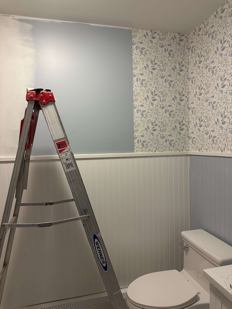 Ladder in front of unpainted white beadboard and a half wallpapered bathroom wall. 